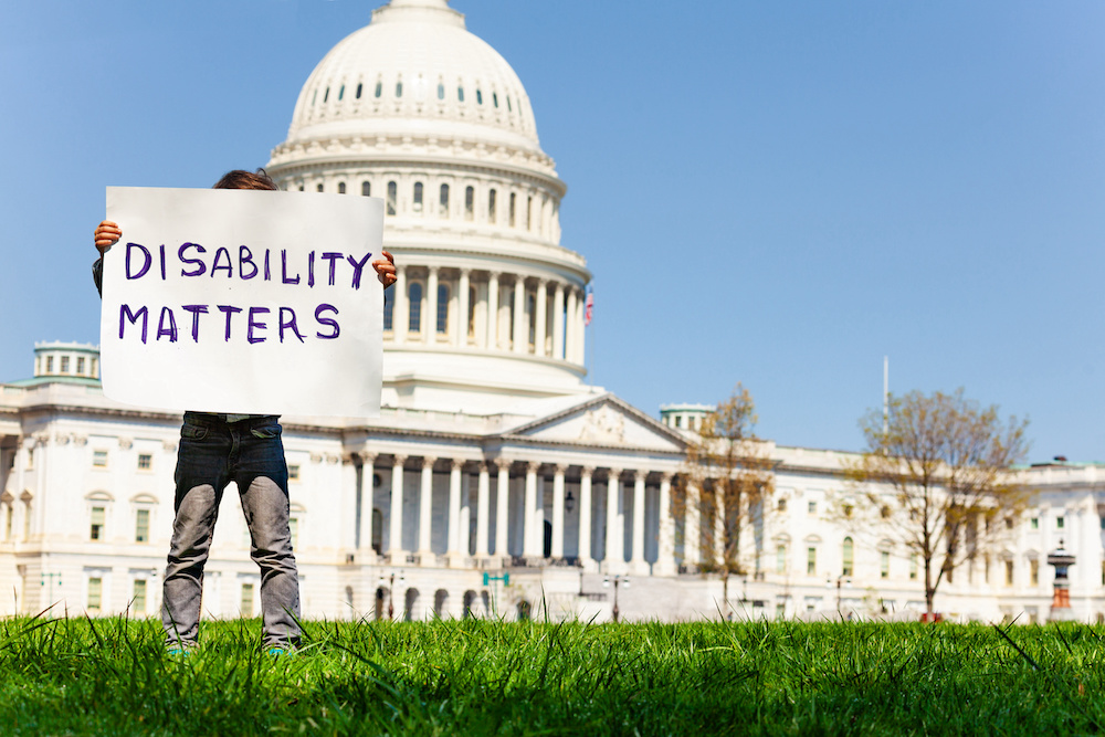 Younger boy standing in front of the USA Capitol building while posing behind a sign that writes "DISABILITY MATTERS."