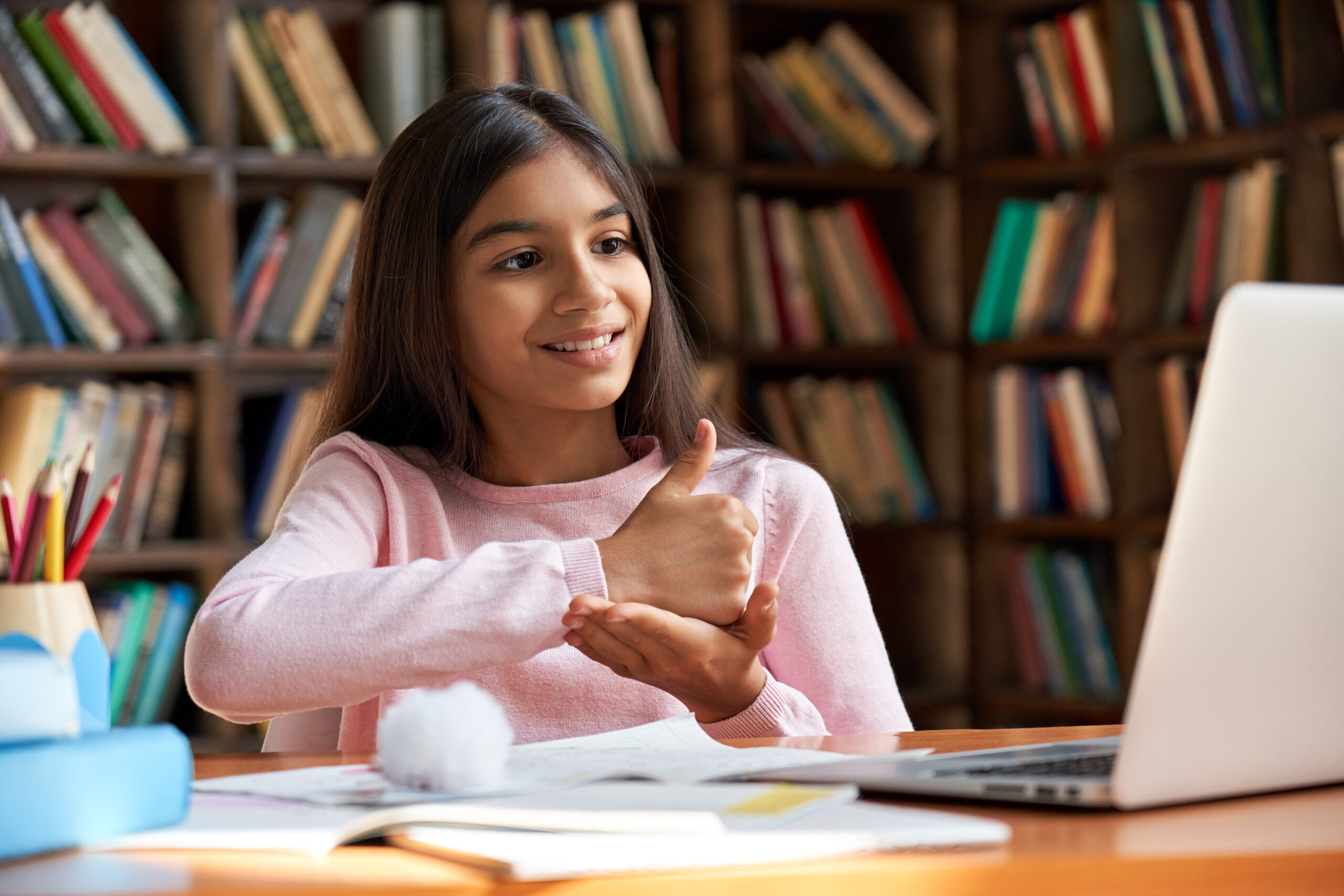 girl sitting in a library smiles and performs sign language directed toward her laptop screen.