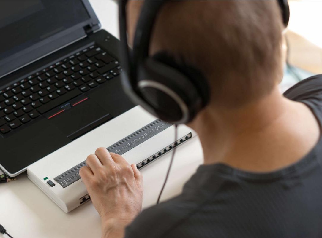 Man wearing headphones touches a brail keyboard set in front of a laptop.
