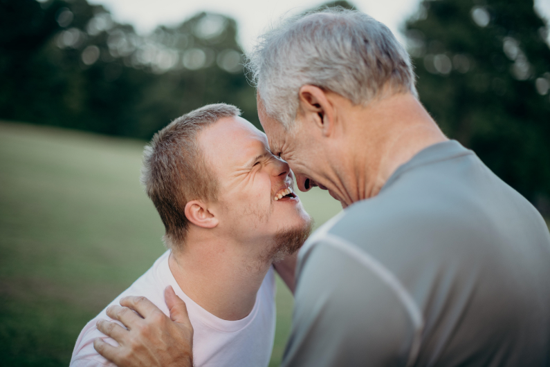 A boy with down syndrome smiles back at an older man holding his shoulders.
