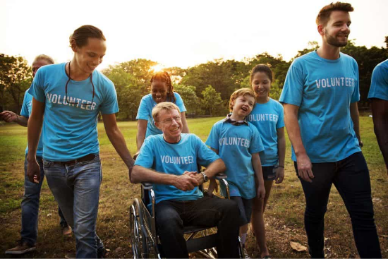 A group of volunteers wearing blue volunteer shirts walk outside. One volunteer is in his wheel chair being pushed a women.